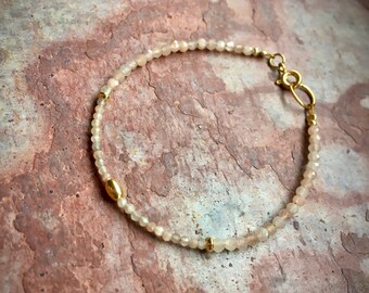 Könnte beinhalten: Ein zartes Armband aus kleinen, hellen Perlen mit goldenen Akzenten. Das Armband hat einen Verschluss.