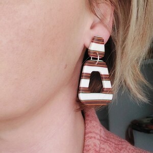 Könnte beinhalten: Ein Paar geometrische Ohrringe mit braunen und weißen Streifen. Die Ohrringe sind aus leichtem Material gefertigt und haben ein modernes Design.