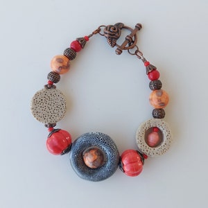 May include: A bracelet with a copper clasp and a rose design. The bracelet is made with orange, red, and gray beads. The beads are made of ceramic and have a textured surface.