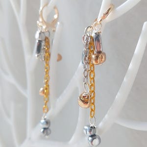 Op de afbeelding: Een paar oorhangers in goud- en zilverkleuren. Elke oorbel heeft een gouden ring, met meerdere kettingen en kralen in goud en zilver. De oorbellen worden gepresenteerd op een witte, abstracte boomachtige structuur.