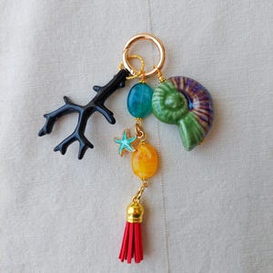 May include: A gold keyring with a black coral charm, teal and yellow beads, a red tassel, and a green and purple nautilus shell charm. The keyring also features a small blue starfish charm.