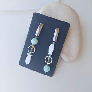 Può includere: Un paio di orecchini pendenti con una parte superiore rettangolare color argento, una perla rotonda azzurra, un piccolo anello color argento e un ciondolo bianco a forma di pesce. Gli orecchini sono esposti su un cartoncino rettangolare blu scuro.