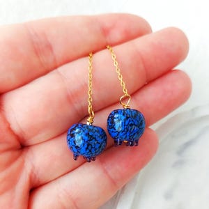 Könnte beinhalten: Ein Paar blau-schwarze Perlenohrringe mit goldfarbenen Ketten. Die runden Ohrringe haben ein strukturiertes Muster und kleine lila Akzente. Sie hängen an einer goldfarbenen Kette.