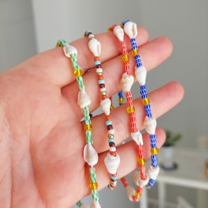 Può includere: Quattro collane di conchiglie con perline in verde, marrone, rosso e blu. Ogni collana presenta piccole conchiglie bianche e perline di vetro colorate. Le collane sono tenute in una mano, mostrando la varietà di colori e design.