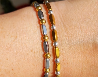 May include: Two beaded bracelets with a mix of gold, silver, and gray metallic beads. The bracelets are worn on a wrist, showcasing the jewelry's design and color variations. The beads are oblong and round.