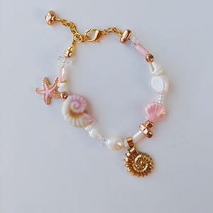 Può includere: Un braccialetto color oro con una varietà di perline, tra cui perle bianche, perline cilindriche rosa e cristalli trasparenti. Il braccialetto è decorato con un ciondolo a forma di stella marina rosa, una conchiglia a spirale e un nautilo dorato.