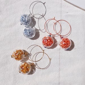 May include: A collection of hoop earrings with clear glass globes filled with silver, red, and gold metallic flakes. The earrings have silver, copper, and gold-colored hoops. The earrings are displayed on a white textured surface.