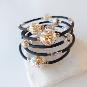 Op de afbeelding: Een zwarte siliconen armband met meerdere strengen en zilveren en gouden vlokken in heldere glazen kralen.