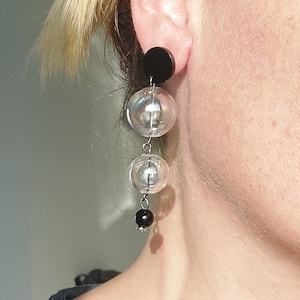 Può includere: Un paio di orecchini pendenti con tre sfere di vetro iridescente e un perno nero in basso. Gli orecchini sono attaccati a un perno nero.