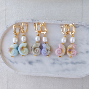 May include: Gold-tone earrings with a seashell design. Each earring features a pearl and a seashell pendant in shades of blue, purple, and pink. The earrings are displayed on a white tray.