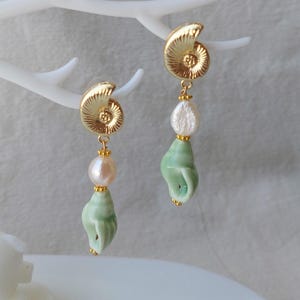Könnte beinhalten: Goldfarbene Ohrringe mit einem Spiraldesign, einer rosa Perle und einem hellgrünen Muschelanhänger. Die Ohrringe hängen an einer weißen, astähnlichen Struktur. Das Design ist zart und feminin, mit Fokus auf natürliche Elemente.