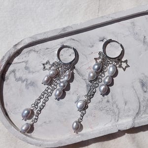 May include: A pair of silver hoop earrings with dangling chains, gray pearls, and star charms. The earrings are displayed on a white and gray marbled tray, showcasing their elegant design and delicate details. The pearls have a lustrous sheen.
