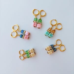 May include: Four pairs of gold hoop earrings with two square-shaped beads hanging from each hoop. The beads are in various colours, including green, blue, pink, and cream.