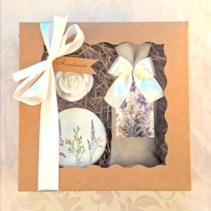 Könnte beinhalten: Geschenkbox aus braunem Karton, verziert mit einem weißen Band und einem Etikett "Handmade". Enthält eine weiße Rose, eine runde Keramik mit Lavendel und einen Jutesack mit Lavendeldruck.