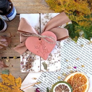 May include: Four rectangular soaps tied with a brown ribbon and a pink heart-shaped tag. The soaps are decorated with dried flowers. The image includes dried citrus slices, autumn leaves, and a bottle.