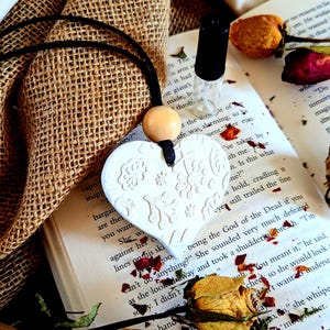 Könnte beinhalten: Ein weißer herzförmiger Anhänger mit floralen Mustern hängt an einer schwarzen Kordel. Eine kleine Glasflasche mit schwarzem Verschluss liegt auf einem offenen Buch mit verstreuten getrockneten Rosenblättern. Der Hintergrund umfasst Jutegewebe und getrocknete Rosen.