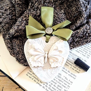 May include: A white heart-shaped ornament with a raised butterfly design and intricate patterns. It has a green ribbon and a small silver button with a tree emblem. The ornament is likely a decorative item or a keepsake.