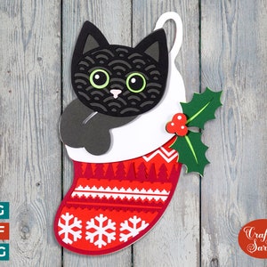 May include: A layered paper Christmas stocking featuring a black cat with green eyes peeking out. The red stocking has a white snowflake and tree pattern. A sprig of holly with red berries is attached. The image includes the text "Craft with Sarah".
