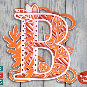 May include: Orange and white layered paper cut out of the letter B with a floral design. The letter is on a woodgrain background. Craft with Sarah is written in cursive at the bottom right corner.
