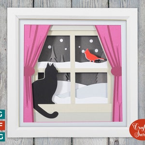 May include: A papercraft window scene featuring a black cat gazing out at a red cardinal perched on a snowy branch. The window is framed with pink curtains and a white frame. The scene is set against a woodgrain background.