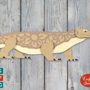 May include: A brown papercraft model of a Komodo dragon, a large lizard native to Indonesia. The model is detailed with intricate cutouts and is posed in a walking position. The background is a woodgrain texture.