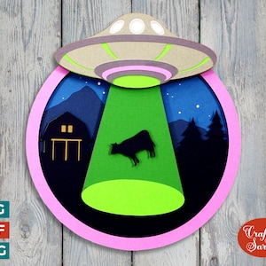 May include: A pink and black paper craft design of a UFO shining a green light on a cow in a field. The UFO is hovering over a farm scene with a barn and trees. The design is on a wooden background.