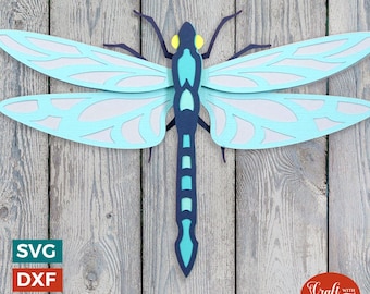 May include: A blue and green paper craft dragonfly with intricate wing details. The dragonfly is against a woodgrain background.