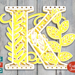 May include: A yellow and white layered papercraft design of the letter K with floral accents. The design is on a woodgrain background with the text "Craft with Sarah" in the lower right corner.