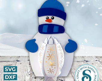 May include: A white snowman figure with a blue hat, scarf, and mittens, holding a white lantern decorated with gold snowflakes. The lantern is open, revealing its interior. The image includes the text "SVG, DXF, PNG" and a circular logo that says "Craft with Sarah. Made with Love."