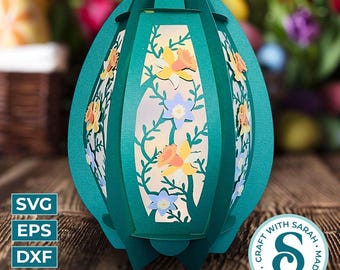 May include: A teal paper lantern in an egg shape, decorated with floral cutouts of yellow, orange, and blue flowers. The lantern has a handle at the top and is sitting on a wooden surface. The image includes the text "SVG, EPS, DXF, PNG" and a circular logo that says "Craft with Love, Made with Sarah."
