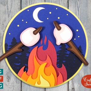 Puede incluir: Un diseño de artesanía en papel de una fogata con dos malvaviscos en palos, sobre un fondo de cielo nocturno azul con una luna creciente y estrellas. El diseño está en un círculo con un borde amarillo.