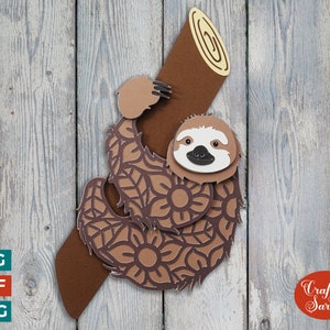 May include: A brown sloth with a mandala pattern hanging from a brown log. The sloth has a white face with black eyes and a black nose. The background is a light wood grain.
