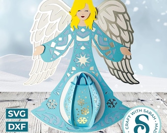 Può includere: Un ornamento a strati a forma di angelo di carta nei toni del blu, bianco e giallo. L'angelo ha le braccia tese, ali dettagliate e una lanterna decorativa. Il design include fiocchi di neve e stelle. Vengono visualizzati anche i formati di file SVG, DXF e PNG.
