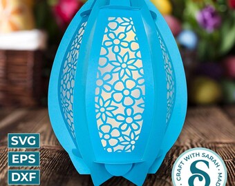 May include: A blue paper Easter egg lantern with a white floral design. The lantern is made of multiple layers of paper and has a light inside. The lantern is sitting on a wooden surface. The text "CRAFT WITH LOVE. MADE WITH SARAH." is on a circle sticker.