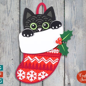 Peut inclure: Un chat noir qui sort d'une chaussette de Noël rouge et blanche avec une branche de houx verte. La chaussette a un motif de flocons de neige blancs et une bordure blanche.