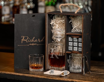 May include: A personalized whiskey decanter set in a wooden box. The set includes a decanter, two glasses, ice stones, tongs, and a gift bag. The decanter and glasses are filled with amber liquid. The box has a rope handle.