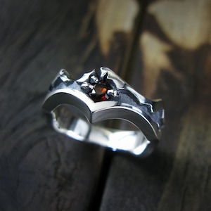 May include: A silver ring with a textured, jagged design. The ring has a single red gemstone in the center.