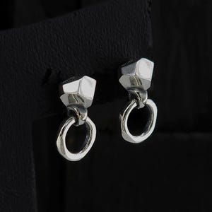Puede incluir: Un par de pendientes de plata. Cada pendiente presenta una parte superior geométrica y facetada conectada a un pequeño anillo abierto. Los pendientes se muestran sobre un fondo oscuro, resaltando su brillo metálico y diseño moderno.