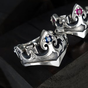 May include: Close-up of two silver crown rings. The rings feature intricate scrollwork and are adorned with blue and red gemstones. The rings have a textured surface and are set against a dark background.