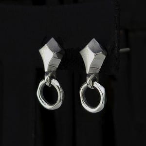 Puede incluir: Un par de pendientes de plata con un diseño geométrico. Cada pendiente presenta una parte superior facetada en forma de diamante conectada a un pequeño anillo texturizado. Los pendientes se muestran sobre un fondo oscuro.