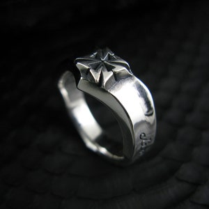 Könnte beinhalten: Ein silberner Ring mit einem Sternmuster auf der Oberseite. Der Ring hat eine polierte Oberfläche und ein dickes Band. Das Sternmuster ist erhaben und hat mehrere Spitzen. Der Ring ist vor einem dunklen Hintergrund.