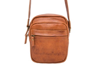 May include: A brown leather crossbody bag with a rectangular shape. The bag has a front zippered pocket, a top compartment, and a long adjustable strap. The bag has a distressed finish and the brand name "Jana Douc" is visible on the bottom.