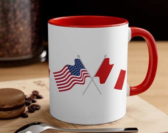 May include: A white ceramic coffee mug with a red rim and handle. The mug features a graphic of a crossed American and Canadian flag.