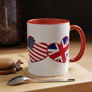 May include: White ceramic mug with a red interior and handle. The mug features two heart-shaped designs, one with the American flag and the other with the British flag. The mug sits on a wooden surface with a spoon and a pastry.