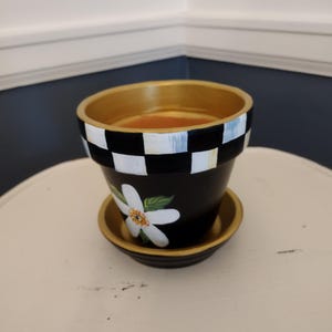 May include: Black and gold painted flower pot with a matching saucer. The pot features a white flower design and a black and white checkered pattern. The rim and saucer have a gold finish. A decorative item for plants.