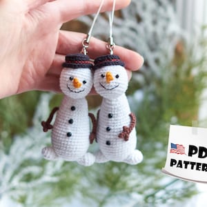 May include: Two crocheted snowman ornaments with black hats and brown accents hanging from silver chains. The ornaments are white with black eyes and orange noses. PDF PATTERN.