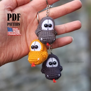 Puede incluir: Tres llaveros de monstruos de ganchillo en gris, amarillo y gris oscuro, cada uno con ojos negros grandes y una lengua roja. Los llaveros están unidos a un llavero plateado. La imagen incluye el texto "PDF PATTERN" y una pequeña bandera americana.