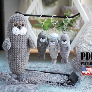 May include: A collection of handmade gray crochet seal keychains and a larger seal plushie. The plushie wears glasses and has a white muzzle. The keychains are hanging from a black bar. The image includes the text "PDF PATTERN".