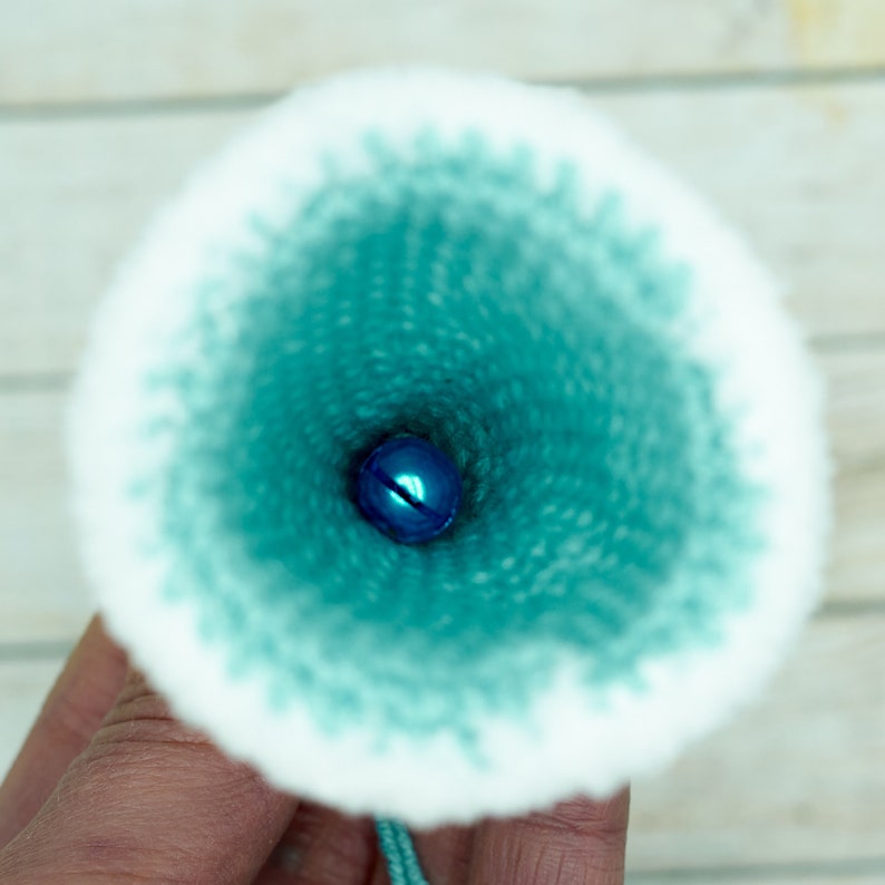 K&ouml;nnte beinhalten: Nahaufnahme des Inneren eines tealblauen, gestrickten Stoffkegels mit einer kleinen blauen Glocke am Boden.