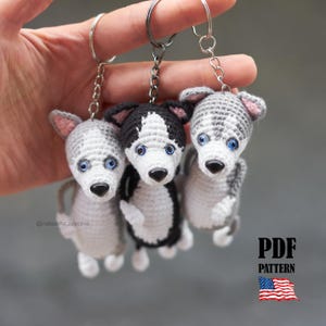 Puede incluir: Tres llaveros de perros husky de ganchillo con ojos azules y narices negras. Los perros son grises, negros y blancos, y están unidos a llaveros plateados. La imagen incluye el texto "PDF PATTERN" y una pequeña bandera americana.